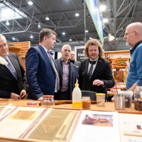 Eine Gruppe an Männern in Anzügen wird von einem anderen Mann an einem Stand beraten. Eine Gruppe an Männern in Anzügen wird von einem anderen Mann an einem Stand beraten.